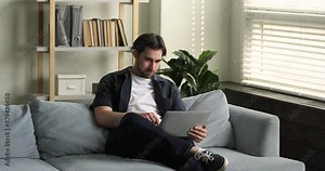 Millennial man sit on sofa at homeoffice texting messages use portable computer, do telecommute job use internet and application, young generation and modern wireless tech usage, distance work concept