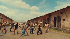 A dynamic group of dancers performing a synchronized routine outdoors, showcasing energy and movement. The setting features rustic buildings and a cloudy sky, enhancing the vibrant atmosphere.