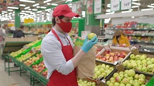 Salesman in red face mask standing in grocery store selecting fresh apples. Courier working in supermarket delivery service during epidemic covid-19, coronavirus pandemic, online food shopping