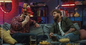 Two Caucasian young guys friends spending their leisure time together in the evening on the couch, one of them wearing beer helmet.