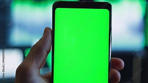 POV pushing towards close up of smartphone held in man's hand where smart phone screen is a chroma key green screen. Background is screen filled with stock trading charts.