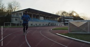 A cinematic shot of young black male running on empty running track with the sun setting in the background.