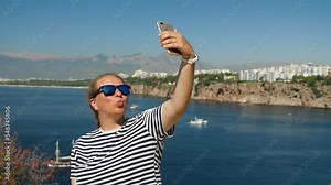Woman making duck lips taking selfies on her phone against the sea. Girl in sunglasses makes duck face taking selfies on her smartphone. Beautiful views in the background