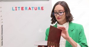 Young woman teacher in glasses opens thick book against whiteboard in classroom. Enjoyable foreign and native literature lessons for pupils at school