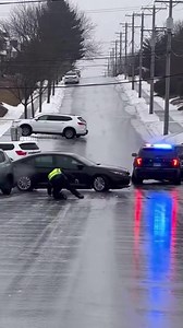 A steep residential street in a neighborhood turned into a dangerous skating rink during a recent ice storm, leading to multiple collisions caught on a bystander's camera. The video shows several cars already damaged at the bottom of the hill when a police officer arrives to assess the situation. The officer slips and falls on the glare ice just as a black sedan begins spinning toward him. The car narrowly misses the fallen officer before crashing into the wreckage. Authorities are urging reside