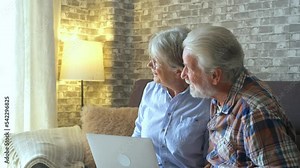 Old couple of two happy pensioners having fun together relaxing on the sofa of beauty and cute house. Portrait of seniors or mature people in love looking at the window.