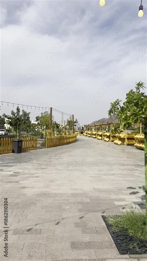 Daytime view of Suq Al-Uyun Market in Medina, near Mount Uhud, featuring yellow wooden stalls, hanging string lights, and modern stone paving, elegantly framed by a wooden fence beneath a cloudy sky.