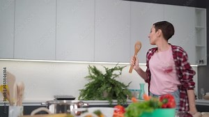 Young happy woman dancing singing using wooden spoon preparing food at new home kitchen, carefree joyful housewife having fun cooking dinner meal listen to music in modern kitchen. FHD footage.