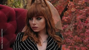 A red-haired model poses in the woods in autumn sitting in a red vintage chair