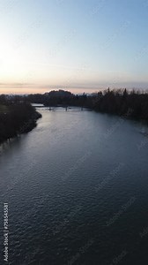 vertical arial over meandering river with water flowing beneath during golden hour, bridge crossing river and forest on either side
