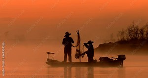 Two silhouetted traditional asian fishermen using nets to fish at an exotic river location from a flat-bottomed boat at sunrise or sunset