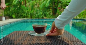 Waiter woman hand serving americano coffee by the swimming pool. Outdoor breakfast on holiday. Asian girl brings cup of caffein drink on table in tropical resort. Travel weekend vacation concept.