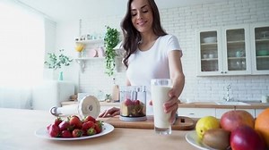 Healthy vegan smoothie with fruits. Beautiful happy young woman in casual home clothes is preparing fitness cocktail with vegetable milk and different season fruits on the kitchen at home. Sporty meal