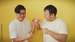 Two asian men laughing and playfully handshaking against an isolated yellow background