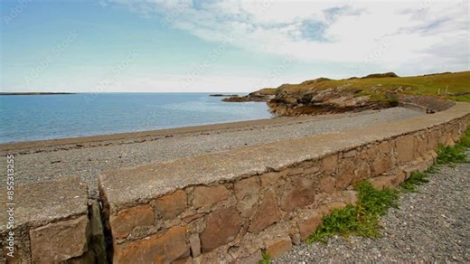 Cemlyn Bay on the North coast of Anglesey, Wales, United Kingdom. Wall, pebble beach and sea, blue sky with rocky shore.