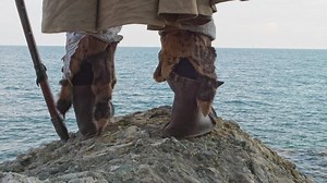 A medieval knight with an ax stands on a stone and looks out to sea. Rear view and closeup of shoes and an axe