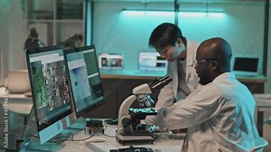 Diverse technical engineers discussing images on computer screen connected to microscope while developing electronics in laboratory