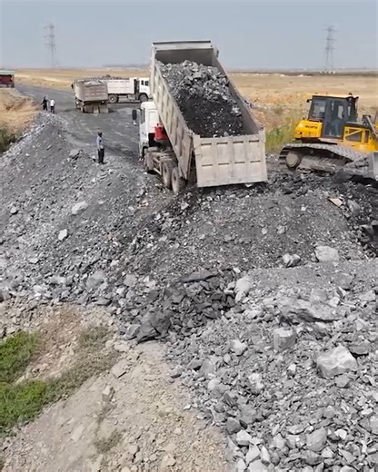 At the edge of the construction site, a skilled bulldozer operator pushes stone while a heavy-duty truck unloads rock for the new road-1 | Excavator Bulldozer
