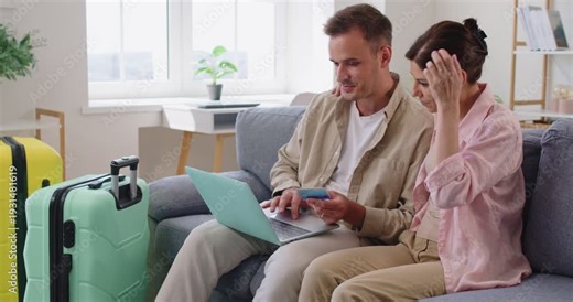 Couple using laptop and credit card to confirm plan of journey and booking, payment before departure. Young happy people sitting together on sofa in home living room, baggage ready for travel