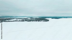Flying over a road on frozen lake ice to reveal a landscape of a snowy forest. Ponds, lakes and woods reach the horizon. Tammela, Finland. Scandinavian nordic landscape on a cloudy day.