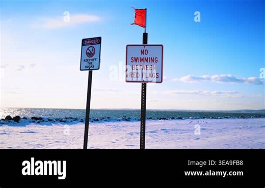 Warning signs on snow covered beach in New York City. Safety signs regarding rocks and swimming near red flag on a snowy shore under clear blue sky Stock Video Footage - Alamy
