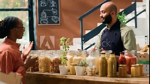 Vendor presents bulk products in jars, recommending type of bio organic pasta to female customer in local grocery store. Young man showing bulk products in recyclable containers, healthy eating.