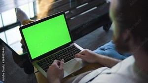 Back view of young man is talking and using laptop while sitting on sofa at home office spbd. Bearded guy hipster having talk and gesturing, looking at computer screen on couch in bright room