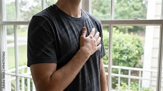 Man placing hand over heart, conveying sincerity, respect, or a pledge indoors by a window with natural light.