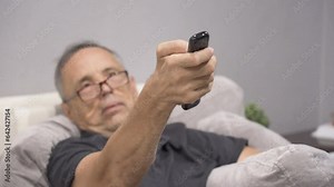 An older man with glasses is looking for his favorite TV program while sitting on the couch