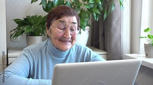 An elderly woman is on a video call, looking at her laptop screen and happily talking. The use of technology for independent living and the importance of online communication for older adults.