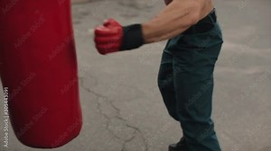 Close side view of muscular male boxer practicing punches and kicks with a punching bag being outdoors. Brown wall on the background