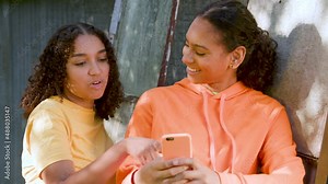 Two beautiful, happy, smiling mixed race African American girls teenagers young women outdoors in the city using a smartphone or cell phone for social media or text messaging
