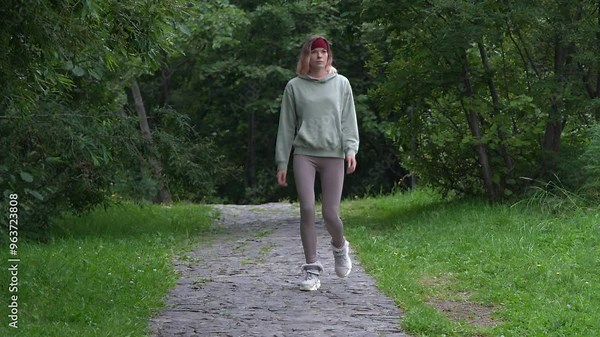Athletic woman dressed in sportswear - tracksuit jacket, leggings and sneakers walking along footpath in park during summer sports training. Full length view, handheld shot, real time video, zoom out