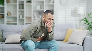 A young adult man is suffering from vomiting and nausea during poisoning while sitting on a sofa at home. A male suffering from indigestion Guy having stomach bug, feeling unwell
