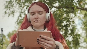 Dreamy pink haired teenage girl in headphones drawing on graphic tablet outdoors