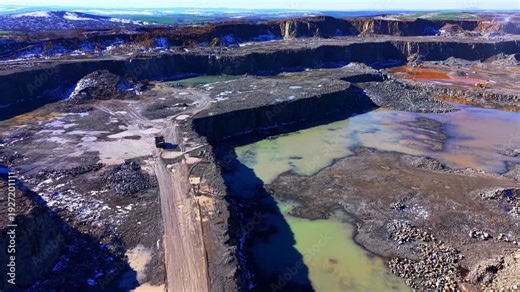 Large scale industrial stone quarry featuring tiered excavation walls, heavy machinery, and pools of mineral-rich water. Wide angle aerial view of open pit stone quarry with water.