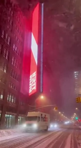 Smartphone footage captures someone standing on a snowy city sidewalk during a blizzard, pointing their phone upward toward a massive digital billboard mounted high on the side of a building. The wind howls loudly through the street, so strong that it completely masks the usual sounds of traffic and the city. Snow whips sideways across the frame as the giant flat screen rattles violently against its mounting brackets. At 0:01 a powerful gust—estimated around 100 mph—catches the back of the billb
