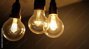 Glowing Edison bulbs hang in darkness. Warm filaments illuminate, creating ambient light. Swinging motion and flickering emphasize fragility and energy dynamics.