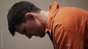 A sad, depressed attractive, handsome man, criminal prisoner, inmate in orange jumpsuit sitting in prison cell thinking, pondering, alone.