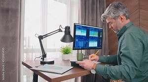 Side view close up portrait of middle-agged broker writing in notepad, computer with graphs on sceen. Trader cheking stock graphs, stock market. 4k. Office work concept.