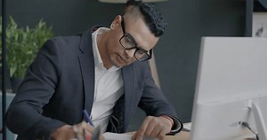 Portrait of Middle Eastern employee typing with PC and writing information in notebook at desk in office. Job and business development concept.