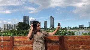 Chinese girl standing outside taking selfies with camera. China woman outdoors shooting self shot with pro camera, posing and mugging. Cute girl takes photos with point and shoot vlog camera.