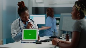 Woman doctor pointing at tablet with horizontal green screen, explaining technology to patient at desk. Medic holding chroma key with mockup template and isolated background on gadget.