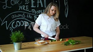 Beautiful young woman cuts green cucumber with kitchen knife on round pieces on wooden cutting board and prepares healthy fresh salad, next to it there green lettuce leaves and tomato with cucumber on