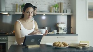 Beautiful Young Woman Sits at the Table in the Kitchen while Uses Tablet Computer, Sips Coffee and Eats Croissants. Young Man in the Background Cooks. Shot on RED Cinema Camera in 4K (UHD).