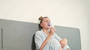 Young woman sitting on bed and putting mask on face to do therapeutic inhalation. Woman inhaling for cough treatment. Nebulizer treatment at home. Self-treatment with saline solution and inhaler
