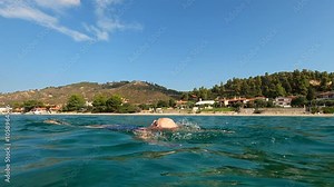 A serene scene depicts an elderly woman swimming in the calm sea near a beach under a clear blue sky, highlighting tranquillity and adventure in nature.