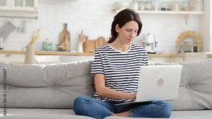 Exhausted young woman suffering from heatstroke flat without air-conditioner, waving blue fan, sitting on couch at home, working on laptop computer. Overheating, high temperature, hot summer weather.
