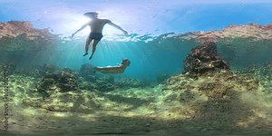 Panorama spherical VR. Two people swim underwater in clear blue water, exploring coral and rocks. Sunlight filters through the surface in a 360 view, creating a stunning immersive scene.