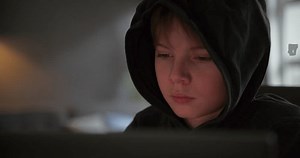 a young boy works on a computer and looks tired. He wears a black hoodie.